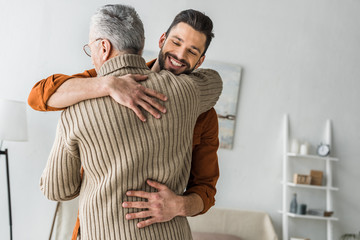 happy bearded man smiling while hugging elder father at home - obrazy, fototapety, plakaty