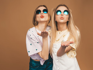 Two young beautiful smiling blond hipster girls in trendy summer colorful T-shirt clothes. Sexy carefree women posing near beige wall in round sunglasses. Positive models giving air kiss