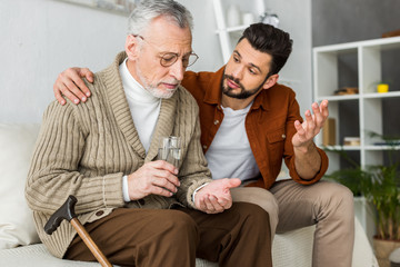 handsome man gesturing senior father holding glass of water and pill