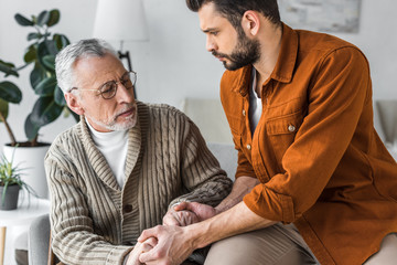senior father in glasses and handsome son holding hands at home