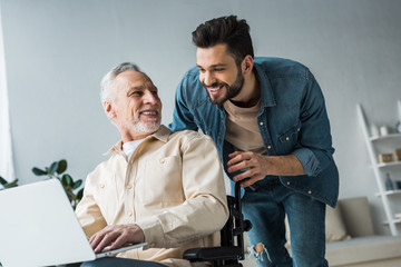 cheerful disabled retired man sitting in wheelchair near handsome son looking at laptop