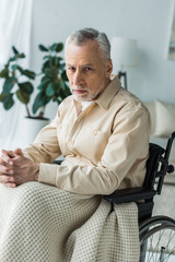 sad disabled retired man sitting in wheelchair with clenched hands and looking at camera