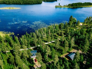 Aerial view of blue lake with green forests in Finland. Wooden house, sauna, boats and fishing pier by the lake.