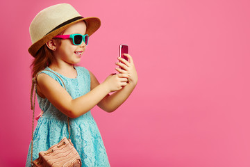 Beautiful little girl with charming smile looks at phone, has a good mood, dressed in fashionable...