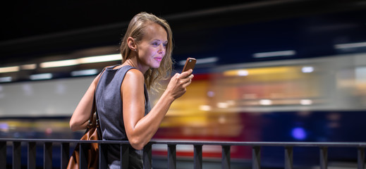 Pretty, young female commuter waiting for her daily train in a modern trainstation, using her cellphone while waiting (color toned image)