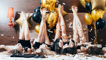 Party women. Fun and joy. Glitter confetti and balloons decor. Young females in black lying on bed legs up. Festive mood.
