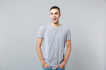 Portrait of excited young man in casual clothes keeping mouth open, holding hands in pockets isolated on grey wall background in studio. People sincere emotions, lifestyle concept. Mock up copy space.