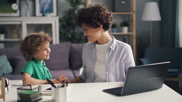 Girl working with laptop at home then talking with her cute little son and kissing him playing with him at home. Motherhood, freelance work and apartment concept.