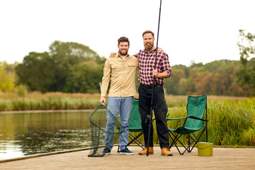 leisure and people concept - happy male friends with fishing rod and scoop net hugging at lake or river
