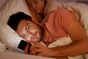 Obraz na płótnie Canvas technology, internet addiction and cheat concept - man using smartphone at night while girlfriend is sleeping