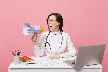 Female doctor sit at desk work on computer with medical document hold megaphone in hospital isolated on pastel pink background. Woman in medical gown glasses stethoscope. Healthcare medicine concept.