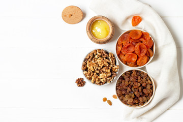 Dried apricots, raisins, walnuts in cups and a barrel of honey standing on a napkin on a white wooden background.