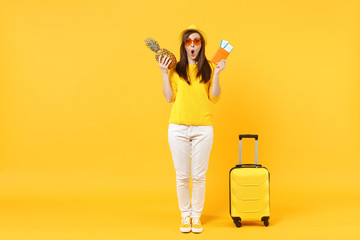 Amazed traveler tourist woman in hat holding passport tickets fresh pineapple fruit isolated on yellow orange background. Passenger traveling abroad on weekends getaway. Air flight journey concept.