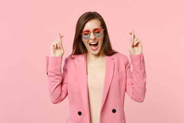 Cheerful young woman in heart glasses waiting for special moment keeping fingers crossed making wish isolated on pastel pink background. People sincere emotions, lifestyle concept. Mock up copy space.