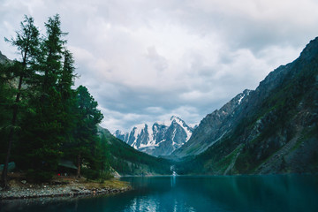 Wonderful mountain lake with view on giant glacier. Amazing huge mountains with conifer forest. Larch tree on water edge. Morning landscape of majestic nature of highlands. Cloudy mountainscape.