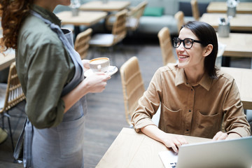 Waitress in apron bringing coffee to businesswoman working with laptop while waiting for order in modern cafe