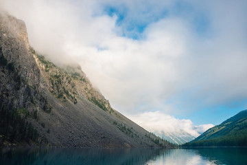 Giant cloud above mountains with trees in sunlight. Amazing mountain lake. Mountain range under blue cloudy dawn sky. Wonderful rocks. Morning landscape of highland nature. Low clouds in sunny light.