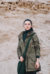 An archaeologist girl in olive-colored clothes stands against a background of sand.