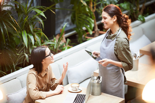 Content confident attractive young lady in glasses sitting at table with laptop and coffee cup and gesturing hand while asking waitress about dessert