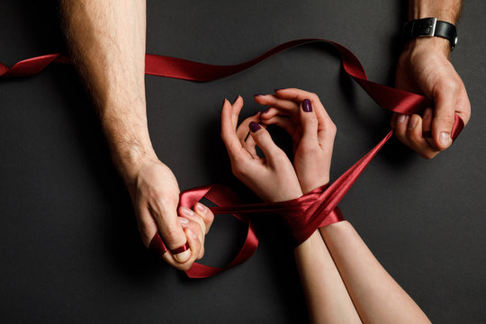 cropped view of man tying red satin ribbon on female hands on black background