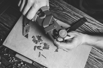 Master holding a hole punch and a piece of leather. On brown wooden table scattered with tools and accessories. - obrazy, fototapety, plakaty