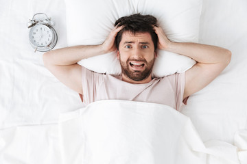 Screaming displeased young man in the morning under blanket in bed lies.