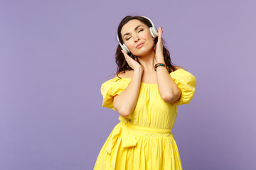 Calmed young woman in yellow dress keeping eyes closed, listen music, putting hands on headphones...