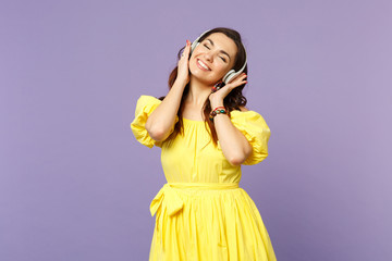 Smiling young woman in yellow dress keeping eyes closed, listen music, putting hands on headphones...