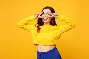 Portrait of laughing young woman in sweater, blue trousers looking camera showing victory sign isolated on yellow orange wall background. People sincere emotions lifestyle concept. Mock up copy space.