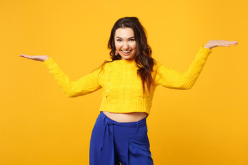 Smiling young woman in sweater, blue trousers looking camera spreading, pointing hands aside isolated on yellow orange wall background. People sincere emotions, lifestyle concept. Mock up copy space.