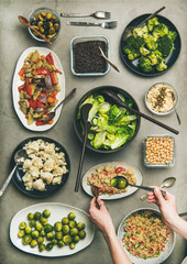 Vegan dinner table setting. Healthy dishes in plates on concrete table. Flat-lay of vegetable salads, legumes, beans, olives, sprouts, hummus and woman hands taking couscous from plate, top view