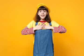 Portrait of amazed girl teenager in french beret and denim sundress keeping hands on chest isolated on yellow wall background in studio. People sincere emotions, lifestyle concept. Mock up copy space.
