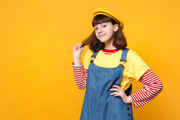 Portrait of smiling cute girl teenager in french beret and denim sundress holding hair isolated on yellow wall background in studio. People sincere emotions, lifestyle concept. Mock up copy space.