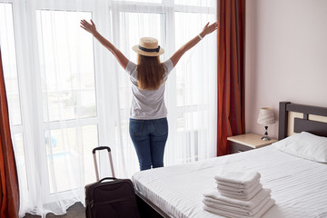 Fototapeta na wymiar Happy young woman staying with raised up arms and luggage in hotel bedroom near big french window, copy space. Back view