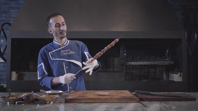 Skillful man in blue chef uniform stringing meat on a skewer, forming lula kebab in modern restaurant. Large grill oven in the background