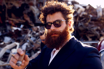Strong bearded man in a classic suit, black sunglasses, smokes a cigar and looks at camera, on iron...