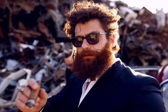 Strong bearded man in a classic suit, black sunglasses, smokes a cigar and looks at camera, on iron dump background.