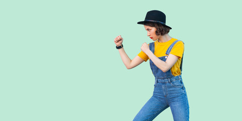 Profile side view portrait of serious young girl in blue denim overalls, yellow shirt, black hat standing with boxing fists and ready to attack. indoor studio shot isolated on light green background