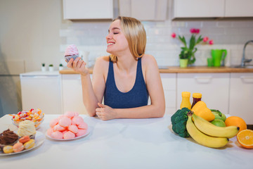 Obraz na płótnie Canvas Young smiling woman with sweet cake in one hand choosing between healthy and unhealthy food in the kitchen. Difficult choice between fresh fruit vegetables or sweets. Dieting. Diet. Healthy Food