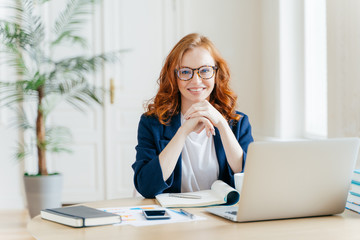 Working concept. Intelligent manager with satisfied expression, writes in notepad, works on modern laptop computer, uses organizer app, sits against office interior. Experienced entrepreneur