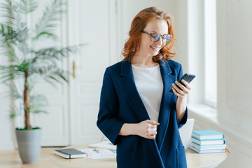 Indoor shot of positive red haired woman checks emial box on modern smartphone device, wears...