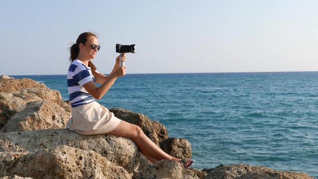 Young woman sitting on stone with camera in hands, capture sea at sunny day