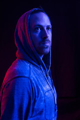 A male model with stubble wearing a hood up in a dark atmospheric future colour setting. Red and blue neon lighting gels. Blade runner neon futuristic man in a black studio.
