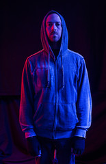 A male model with stubble wearing a hood up in a dark atmospheric future colour setting. Red and blue neon lighting gels. Blade runner neon futuristic man in a black studio.