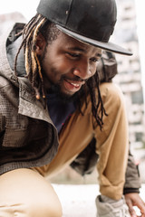 Handsome African American with dreadlocks in brown trousers and a blue sweater in a black cap on his head against the background of houses, city