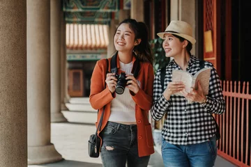 Foto op Canvas two girls tourist visiting bangkok maining attractions in thailand. women friends travel walking in corridor hallway in chinese temple on sunny day in spring. backpackers smiling with camera and book © PR Image Factory