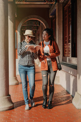 full length asian women travelers searching direction on location map. young girls tourists with camera reading introduction information on guide book walking in corridor of chinese temple sunny day