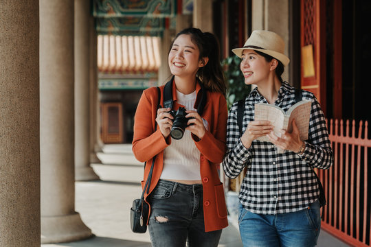 two girls tourist visiting bangkok maining attractions in thailand. women friends travel walking in corridor hallway in chinese temple on sunny day in spring. backpackers smiling with camera and book
