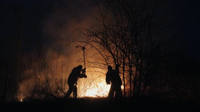Two firefighters with fire flappers extinguish a fire in the forest at night
