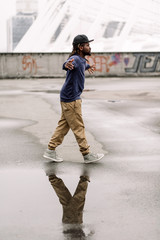 Handsome African American with dreadlocks in brown trousers and a blue sweater in a black cap on his head against the background of houses, city